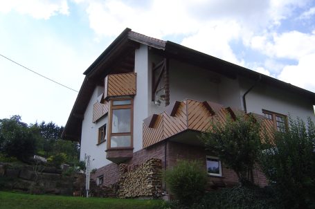 Modernes Haus mit Balkon und Holzverkleidung, umgeben von Bäumen und Wiese.