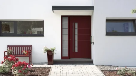 Eingangstür in Rot mit moderner Gestaltung, daneben eine rote Bank und Blumen.