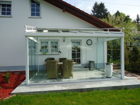 Veranda mit Glaswänden, Stühlen und Tisch, umgeben von grünem Rasen.