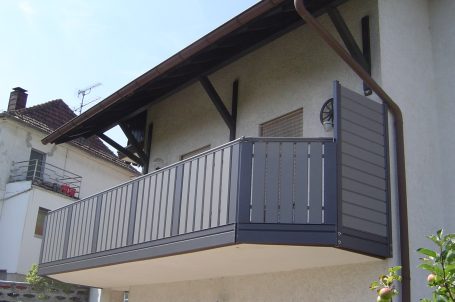 Balkon eines Hauses mit grauer Brüstung und Sonnenschein im Hintergrund.