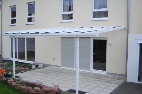 Überdachte Terrasse mit weißen Pfosten und Fliesenboden hinter einem Wohnhaus.