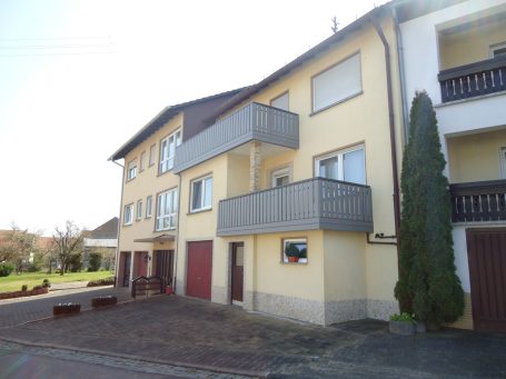 Mehrfamilienhaus mit Balkonen und Garagentoren, umgeben von einem Platz.