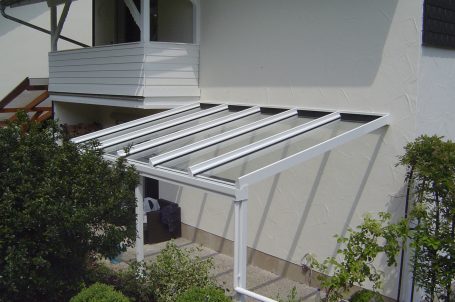 Weißes Terrassendach aus Glas über einem Eingangsbereich mit Schattenwurf.