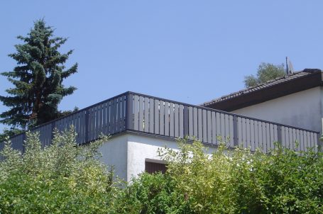 Haus mit Dachterrasse und Holzgeländer, umgeben von Bäumen und blauem Himmel.