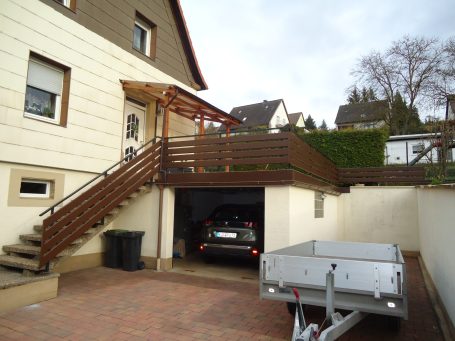 Einfamilienhaus mit Holzterrasse, Garagenauffahrt und Anhänger im Vordergrund.