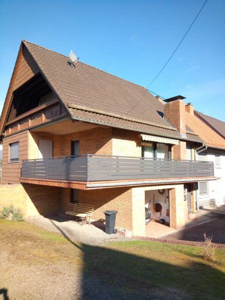 Helles Haus mit Balkon und grauen Fassaden, umgeben von Wiese und anderen Gebäuden.